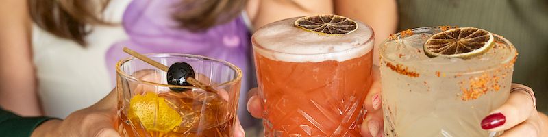 Two women clink drinks&mdash;orange and creamy cocktails with garnishes&mdash;while sharing a plate with a burger and fries at a table.