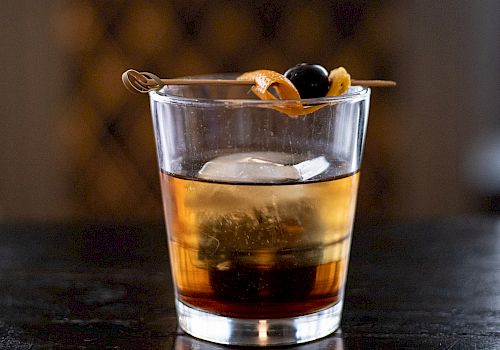 A glass of whiskey on a dark bar, with a twist of citrus peel garnish resting on top.