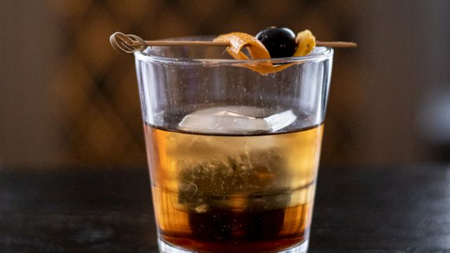 A glass of whiskey on a dark bar, with a twist of citrus peel garnish resting on top.