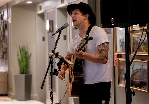 A man in a hat performs guitar and sings into a microphone on stage in a hallway-like venue.