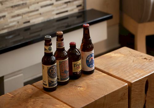 Four beer bottles sit on a wooden bar table by a window, in a cozy pub-like setting.