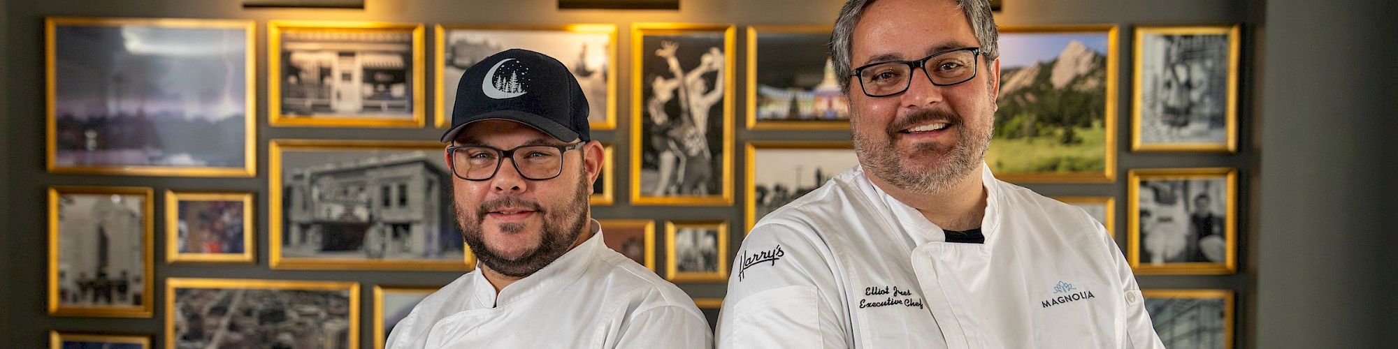 Two chefs in white jackets pose together indoors before a wall of framed photos.
