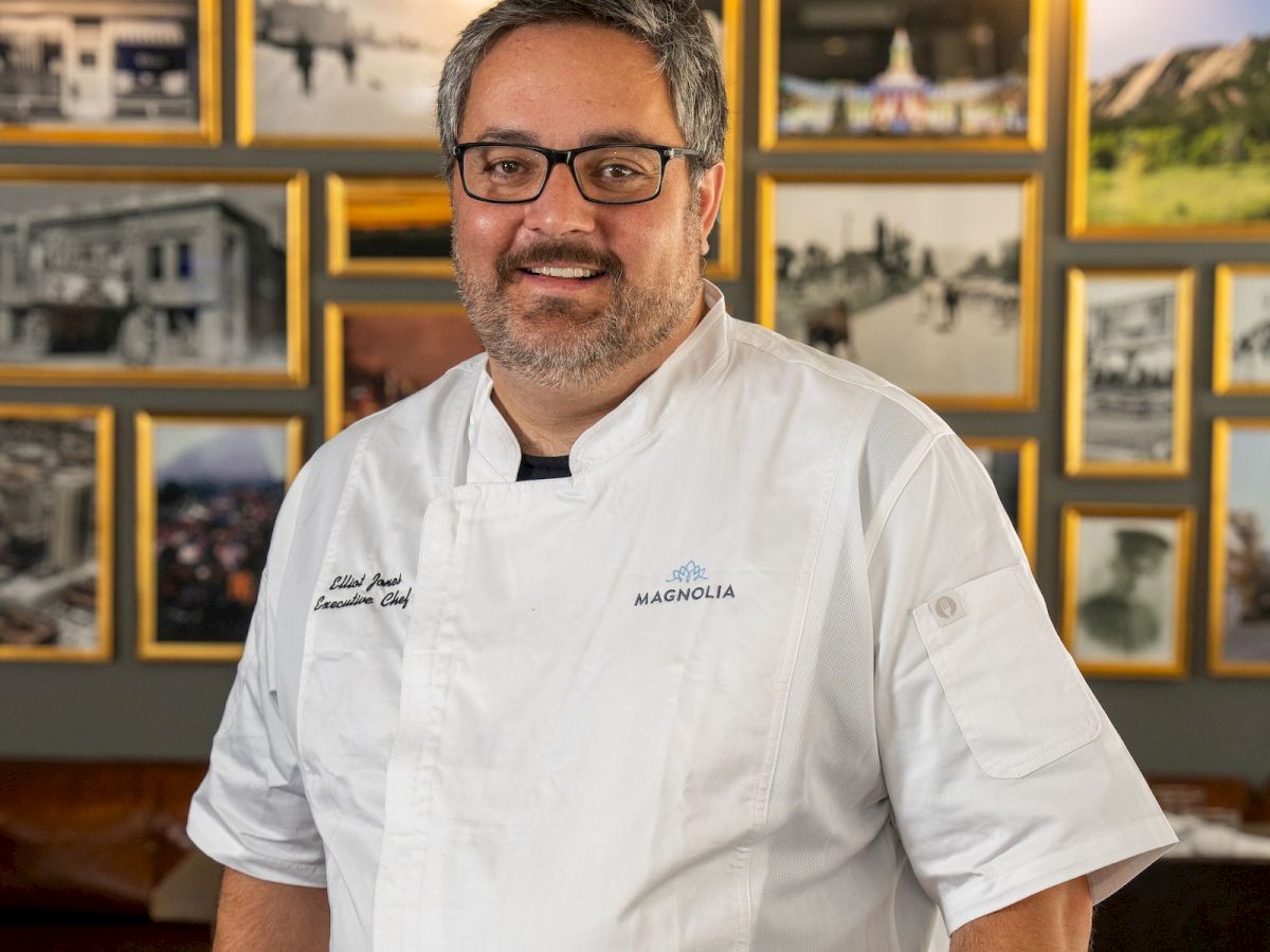 A smiling chef in a white &ldquo;Magnolia&rdquo; coat stands in a restaurant, with framed photos on the wall behind him.