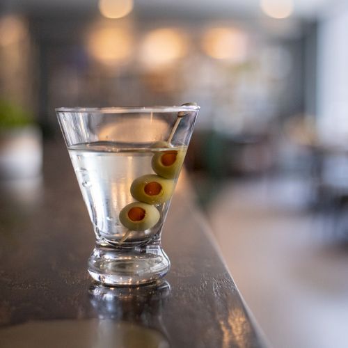 A clear cocktail glass sits on a bar counter, filled with a drink and garnished with green olives.