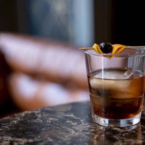 A clear cocktail in a glass, topped with a black olive and a yellow citrus peel on a toothpick.