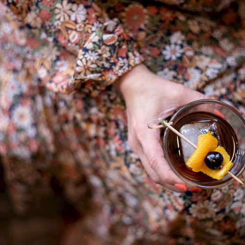 A hand holds a small glass of dark drink with a yellow sponge-cake garnish and a black olive-like piece on a skewer, outdoors.