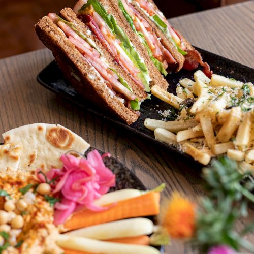 A deli sandwich with stacked cold cuts, greens, and fries on a dark plate, served with fresh salad garnish.
