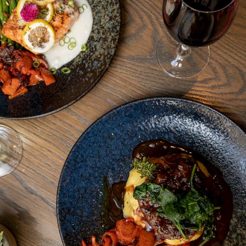A rustic table shows plated meat with roasted veggies, herb garnish, and a glass of red wine.