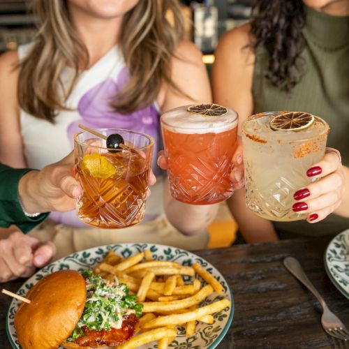 Two friends clink three cocktails on a table with burgers and fries in front.
