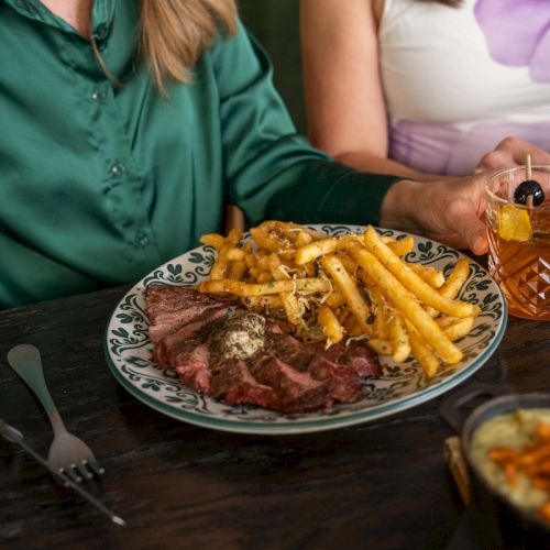 A plate of steak and fries sits on a table while diners sip drinks at a restaurant.
