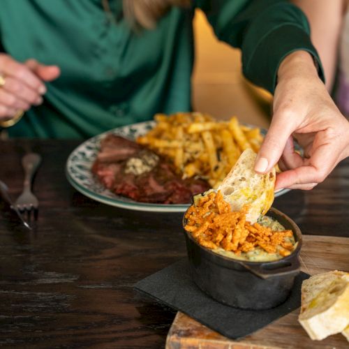 A woman scoops cheesy, spiced food onto bread at a table with fries and steak in the background.