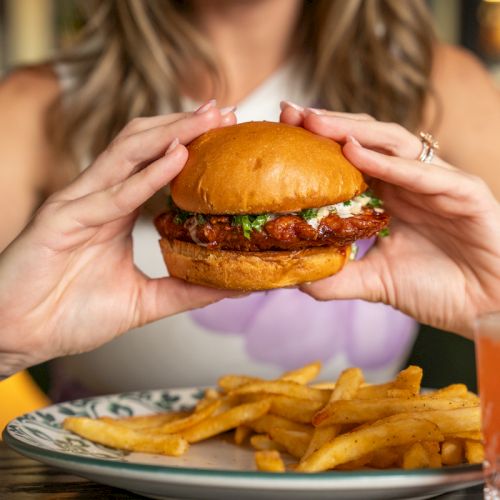 A woman holds a juicy burger with bacon and greens above a plate of fries, smiling in a warm restaurant setting.