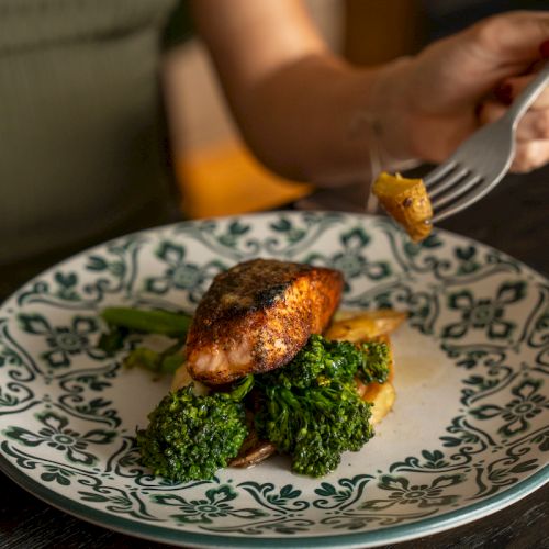 A fork lifts a piece of seared meat from a plate topped with broccoli and potatoes, ready to be eaten.