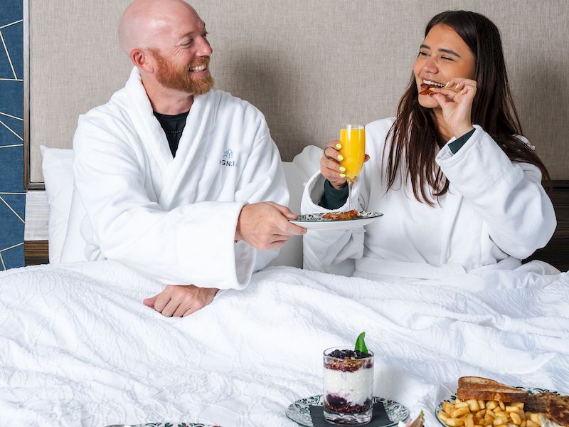A couple in white robes eats breakfast in bed, laughing over plates and juice, with desserts on the table.