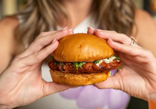 A woman holds a juicy burger with fries on a plate, smiling in a restaurant setting.