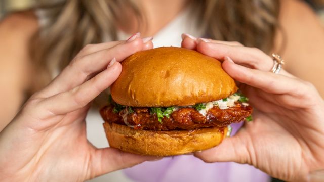 A woman holds a juicy burger with fries on a plate, smiling in a restaurant setting.