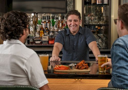 In a cozy bar, a bartender smiles as friends sit at the counter with beer and food, with liquor bottles lining the wall.