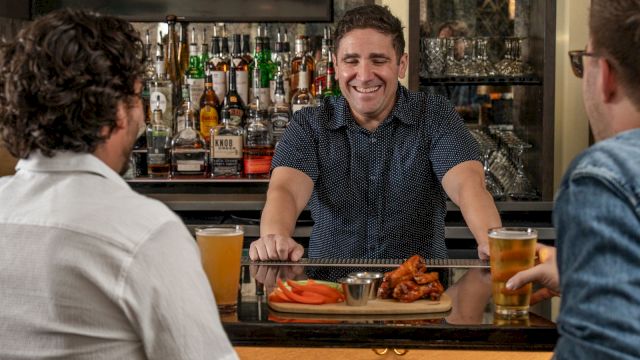 In a cozy bar, a bartender smiles as friends sit at the counter with beer and food, with liquor bottles lining the wall.
