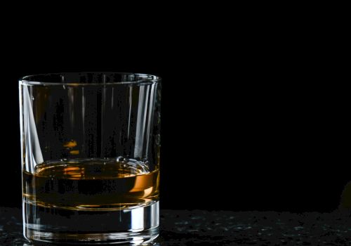 A clear glass sits on a dark surface, filled with amber whiskey (or whisky), with reflections visible in the liquid.