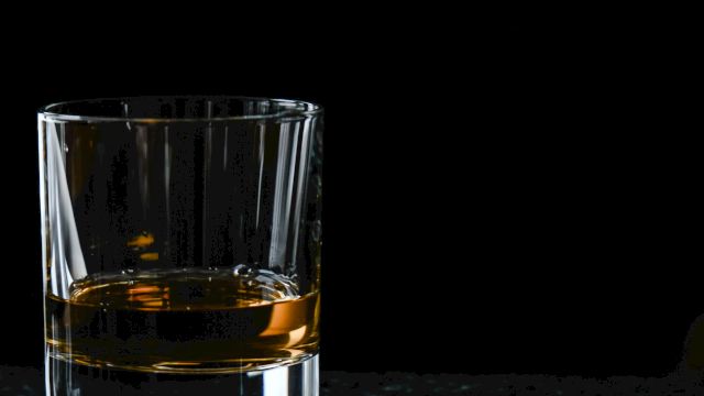 A clear glass sits on a dark surface, filled with amber whiskey (or whisky), with reflections visible in the liquid.