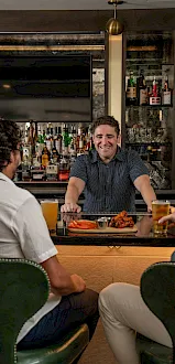 A bartender smiles behind a stocked bar as friends chat over drinks and food, with buffalo wings and carrots on the table.